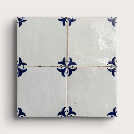 Four square zellige tiles arranged in a 2x2 grid, featuring a glossy off-white glaze with a classic blue floral pattern at the corners and center intersections, set against a light neutral background.