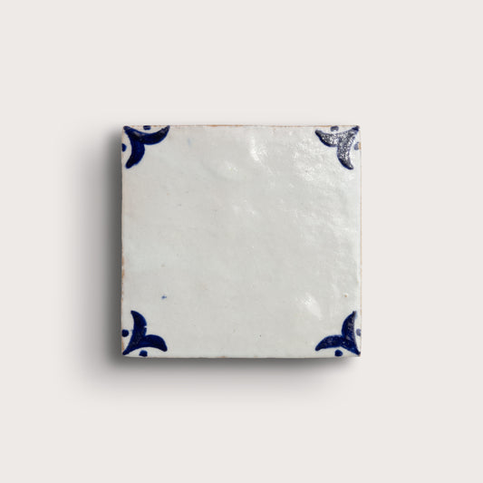 A square zellige tile with a glossy off-white glaze and dark blue decorative corner motifs, shown on a light beige background.