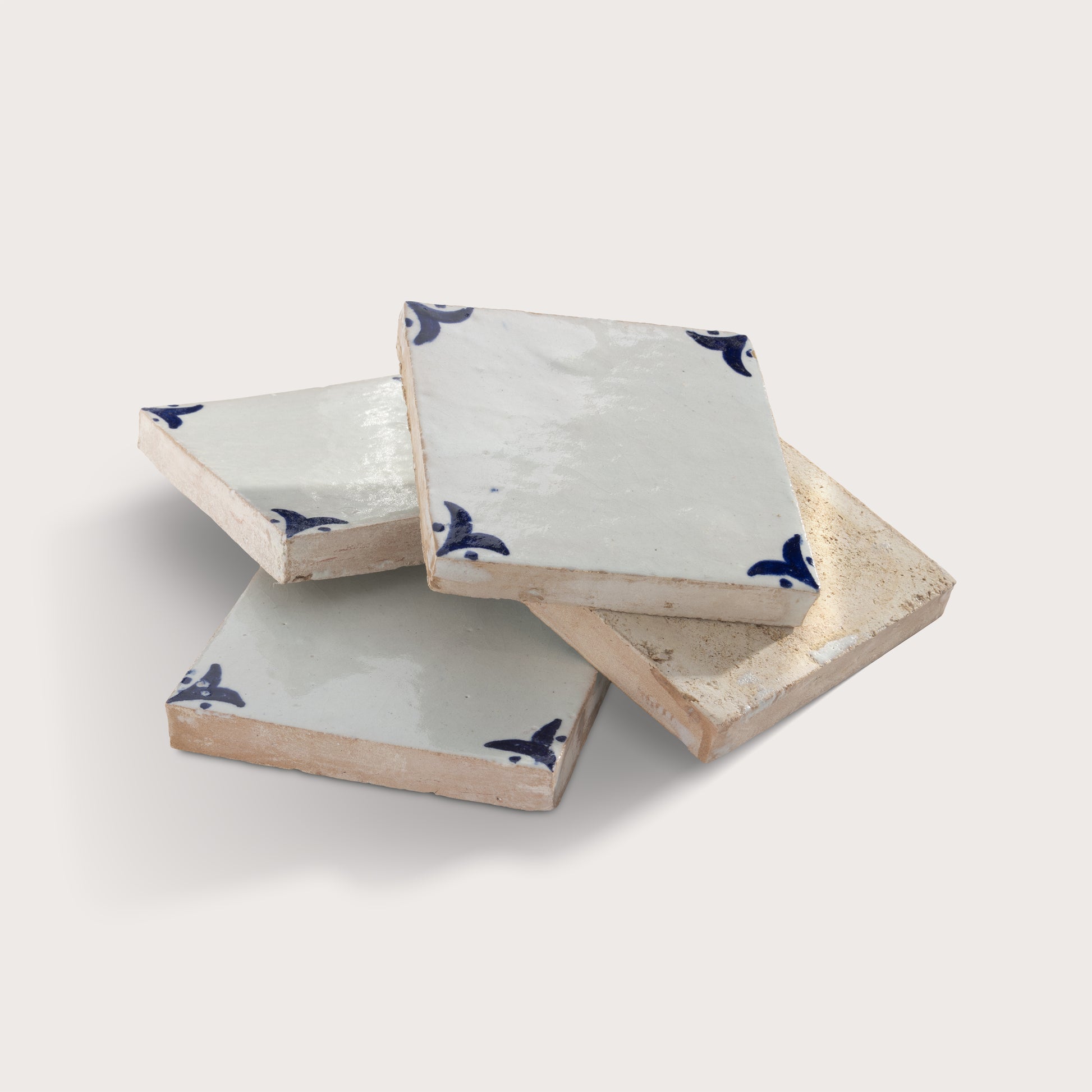 Four square zellige tile stacked in a pile on a light beige background. The coasters have a glossy off-white glazed top with a dark blue bird motif in the corners, and a textured, unglazed beige edge.