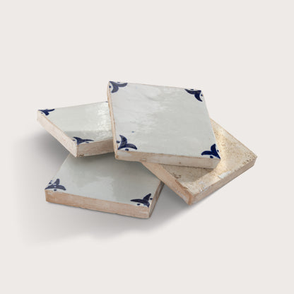 Four square zellige tile stacked in a pile on a light beige background. The coasters have a glossy off-white glazed top with a dark blue bird motif in the corners, and a textured, unglazed beige edge.