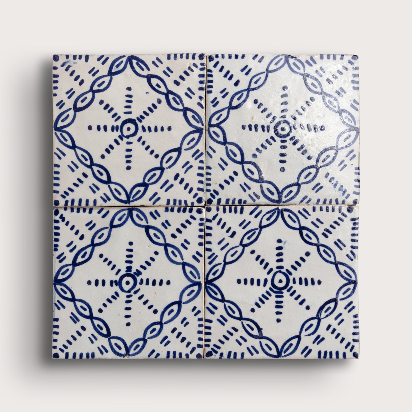 Four square zellige tiles arranged in a 2x2 grid, featuring a blue hand-painted geometric and floral pattern on a glossy white background, set against a light beige wall.