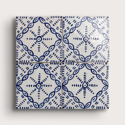 Four square zellige tiles arranged in a 2x2 grid, featuring a blue hand-painted geometric and floral pattern on a glossy white background, set against a light beige wall.