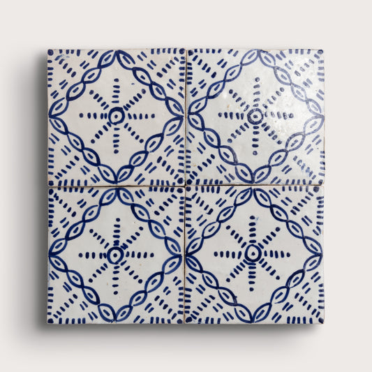 Four square zellige tiles arranged in a 2x2 grid, featuring a blue hand-painted geometric and floral pattern on a glossy white background, set against a light beige wall.