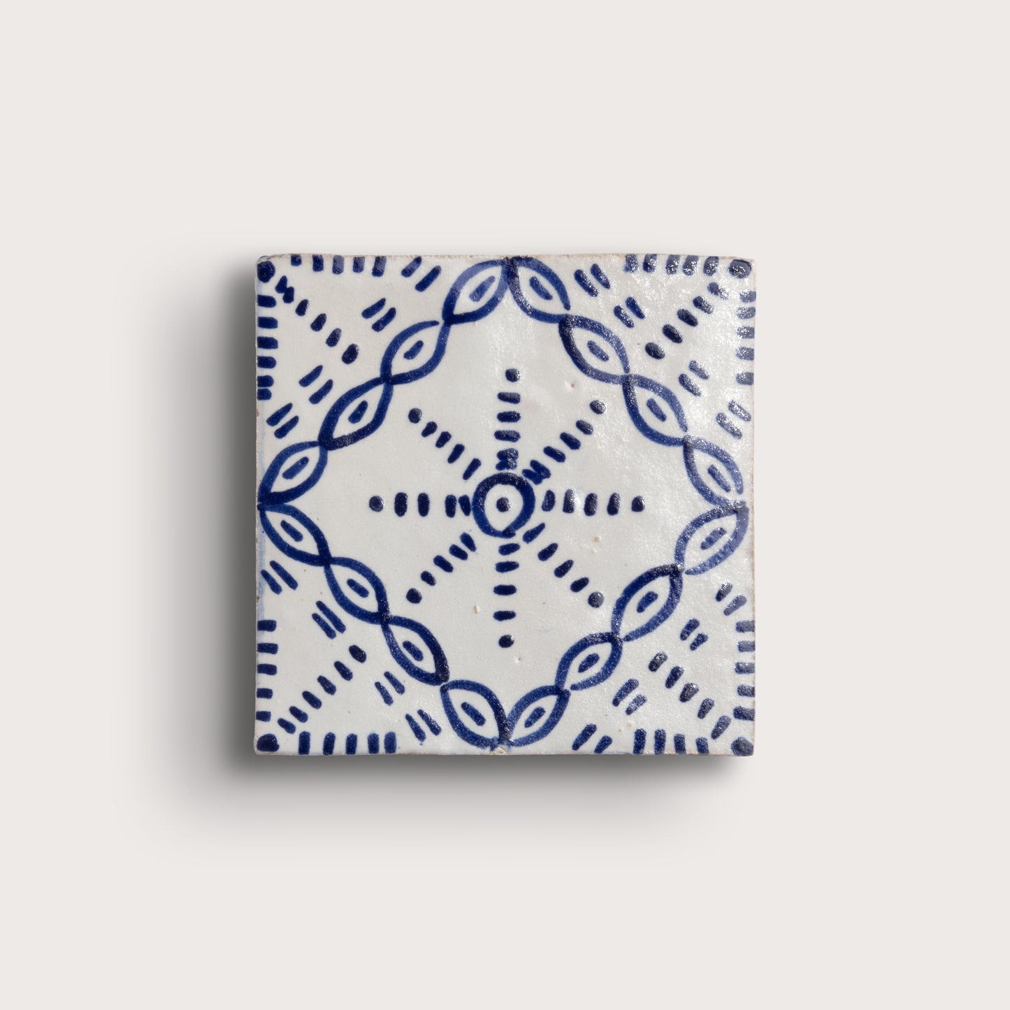 A square zellige tile with a white glaze and a blue hand-painted geometric pattern, shown against a light beige background.