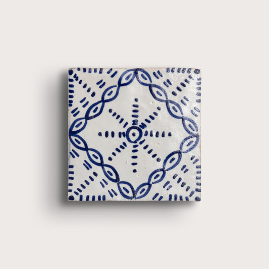 A square zellige tile with a white glaze and a blue hand-painted geometric pattern, shown against a light beige background.