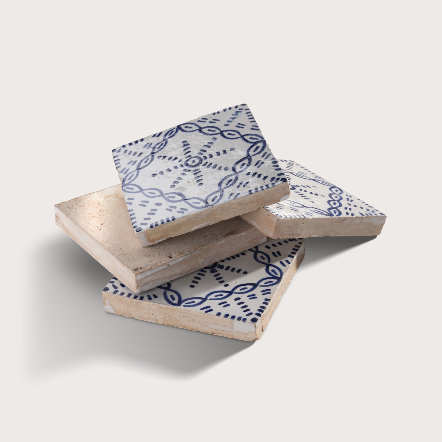 Three square zellige tiles stacked in a pile on a light beige background. The top and bottom tiles feature a white glaze with a dark blue geometric pattern, while the top tile also has a light beige woven or textured inlay visible on its upper surface.