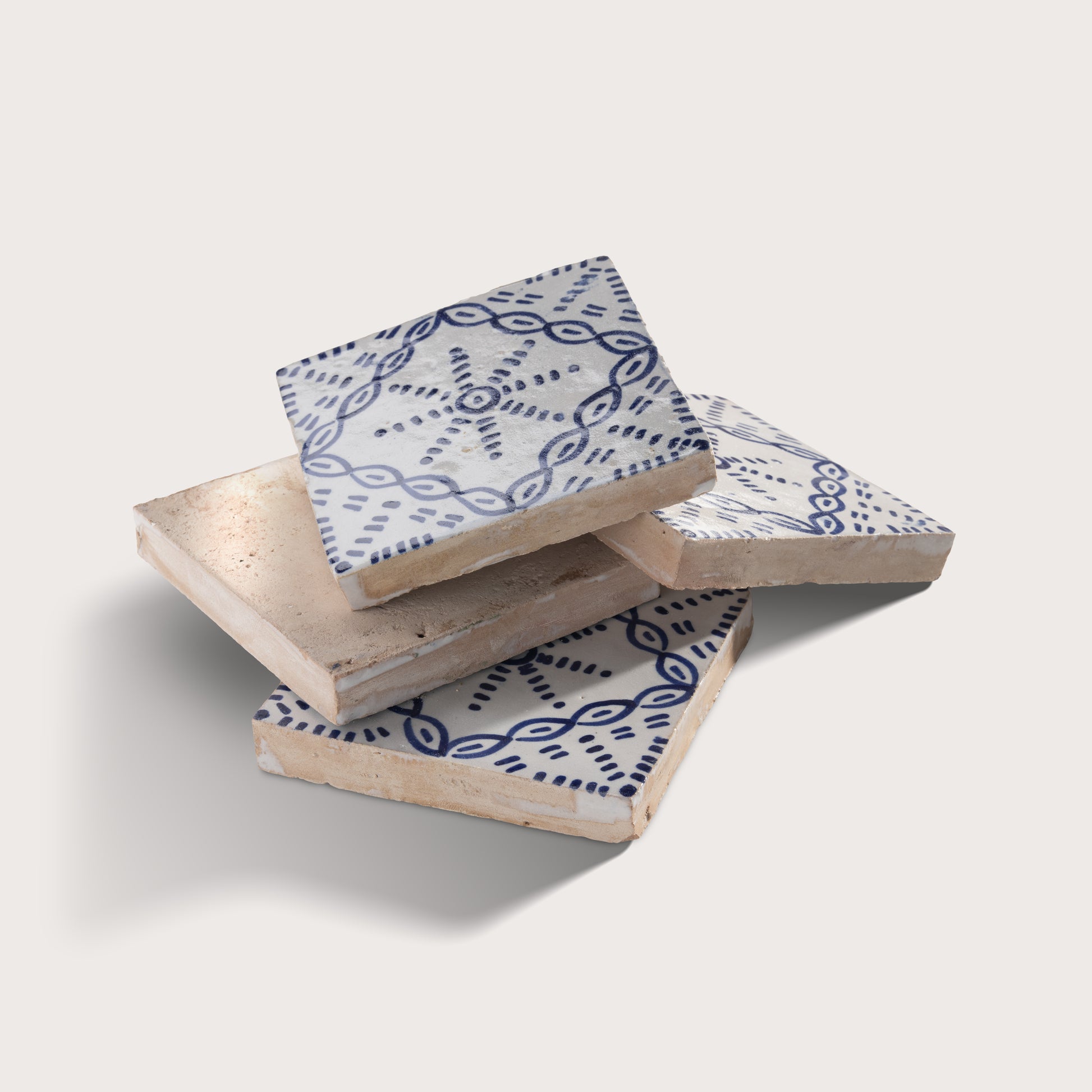Three square zellige tiles stacked in a pile on a light beige background. The top and bottom tiles feature a white glaze with a dark blue geometric pattern, while the top tile also has a light beige woven or textured inlay visible on its upper surface.