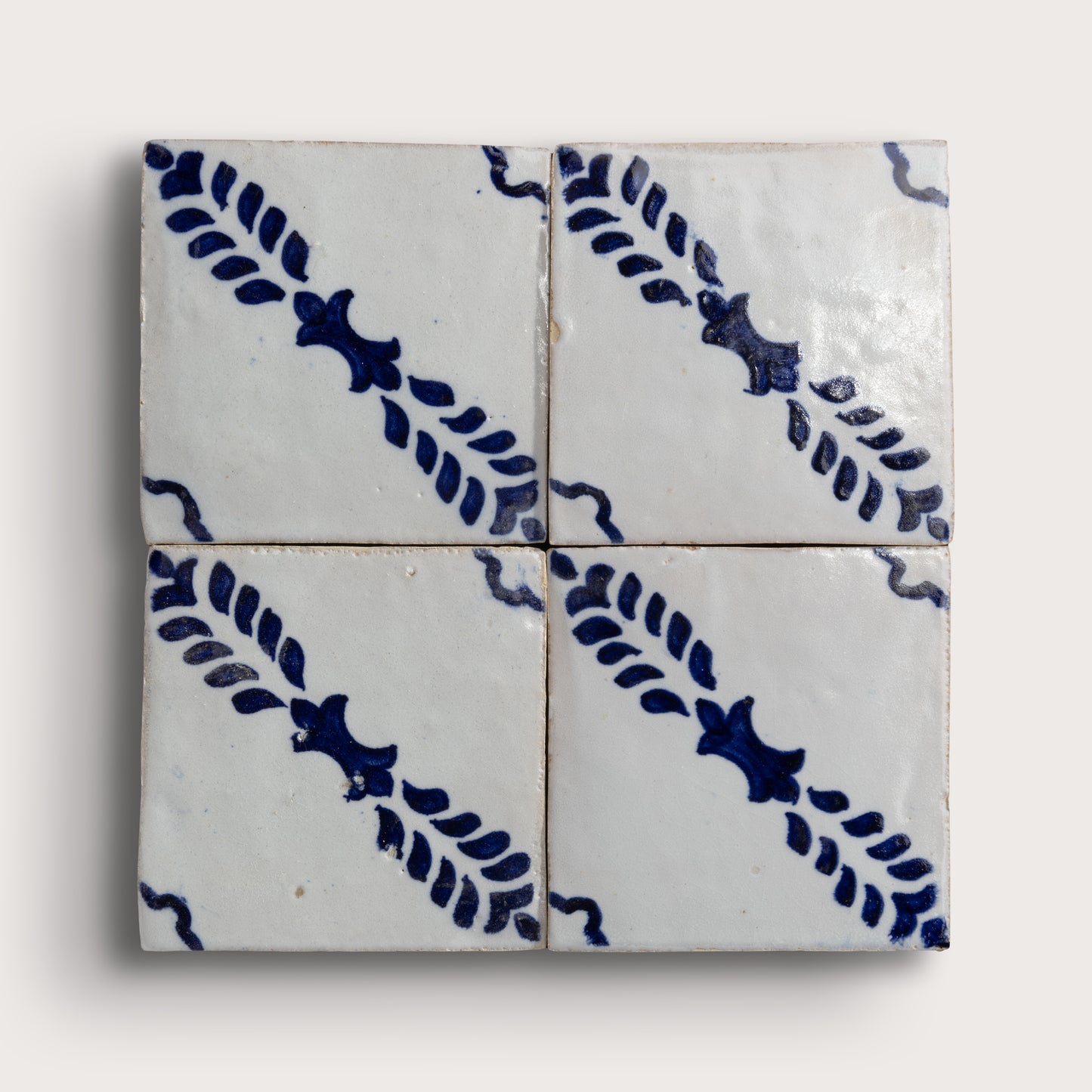 Four square zellige tiles arranged in a 2x2 grid, featuring a white glaze with a dark blue hand-painted diagonal leaf pattern on a light gray background.
