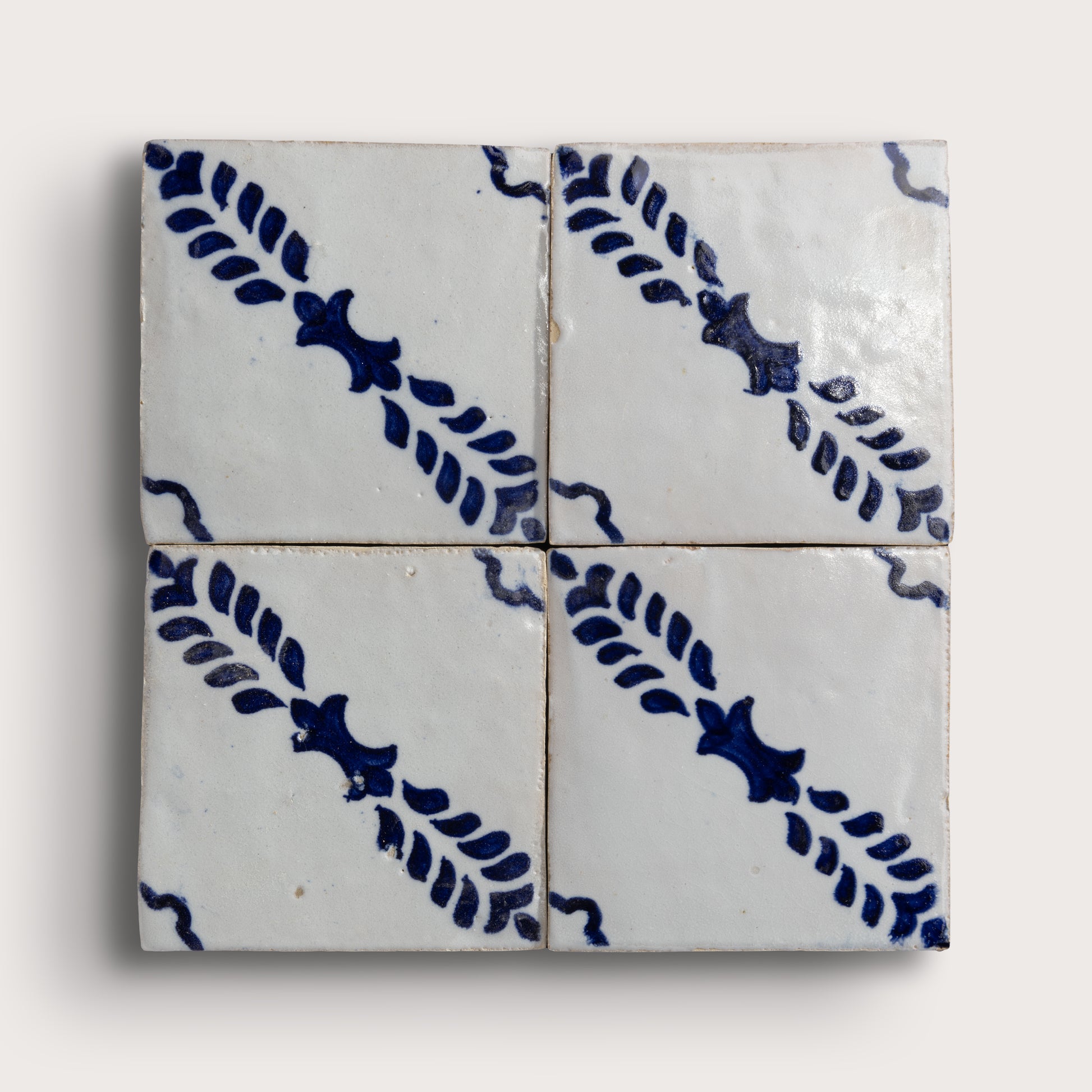 Four square zellige tiles arranged in a 2x2 grid, featuring a white glaze with a dark blue hand-painted diagonal leaf pattern on a light gray background.