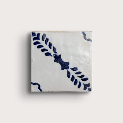 A square white zellige tile with a blue hand-painted floral and leaf pattern, shown against a light beige background.