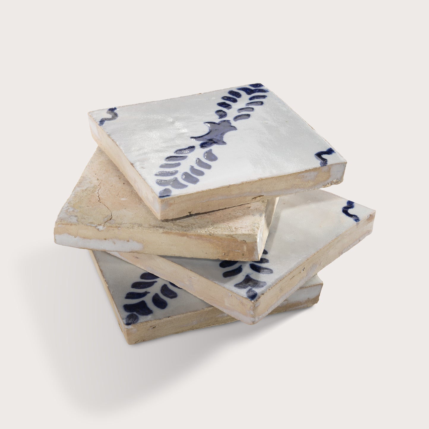 A stack of four rustic zellige tiles with a white glaze, featuring a dark blue painted leaf pattern and showing the natural beige clay edges.