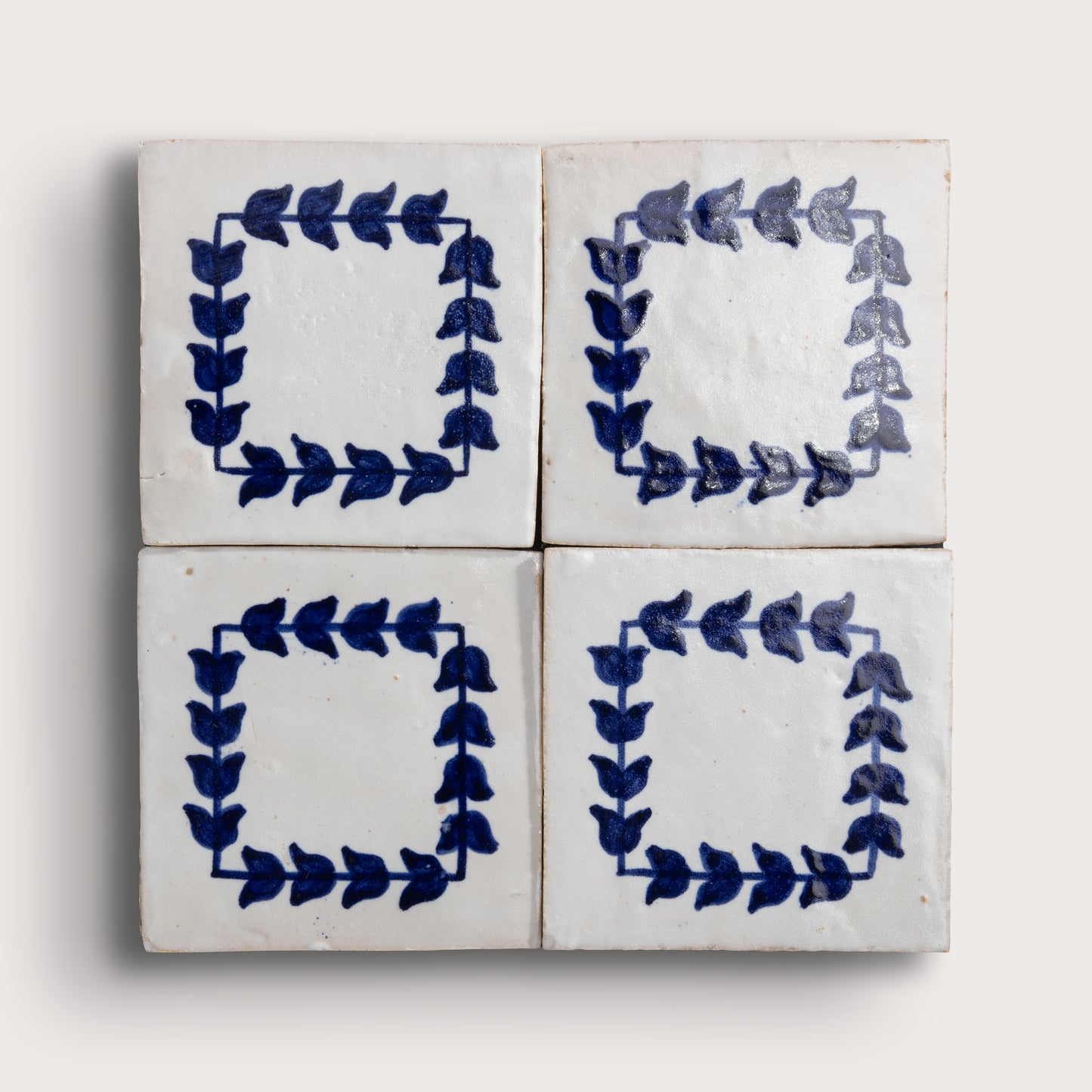 Four square zellige tiles arranged in a 2x2 grid, each decorated with a hand-painted blue leafy border on a white glazed background, set against a light beige wall.