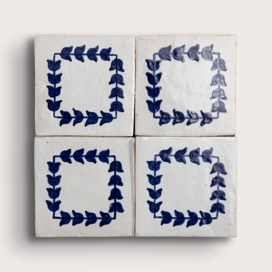 Four square zellige tiles arranged in a 2x2 grid, each decorated with a hand-painted blue leafy border on a white glazed background, set against a light beige wall.