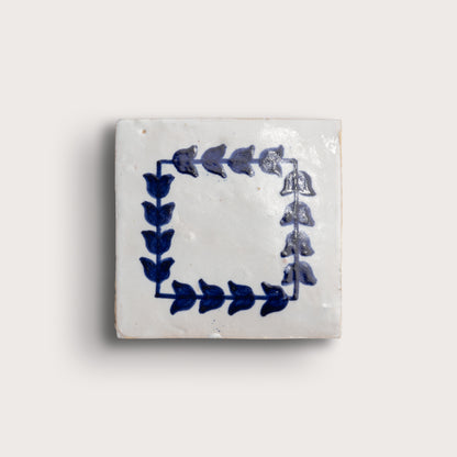 A square zellige tile with a white glaze and a blue floral border design, shown against a light beige background.