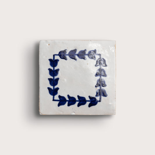 A square zellige tile with a white glaze and a blue floral border design, shown against a light beige background.