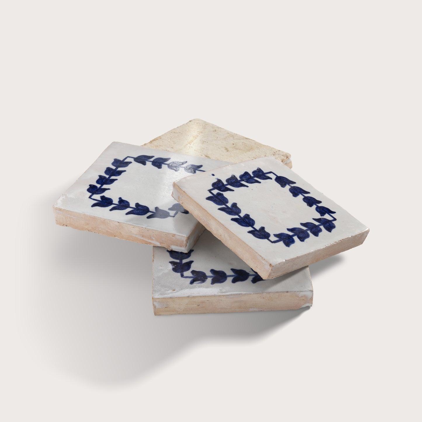 Three square zellige tiles stacked in a fan arrangement, featuring a white glaze with a navy blue leaf border design and a natural beige clay edge.