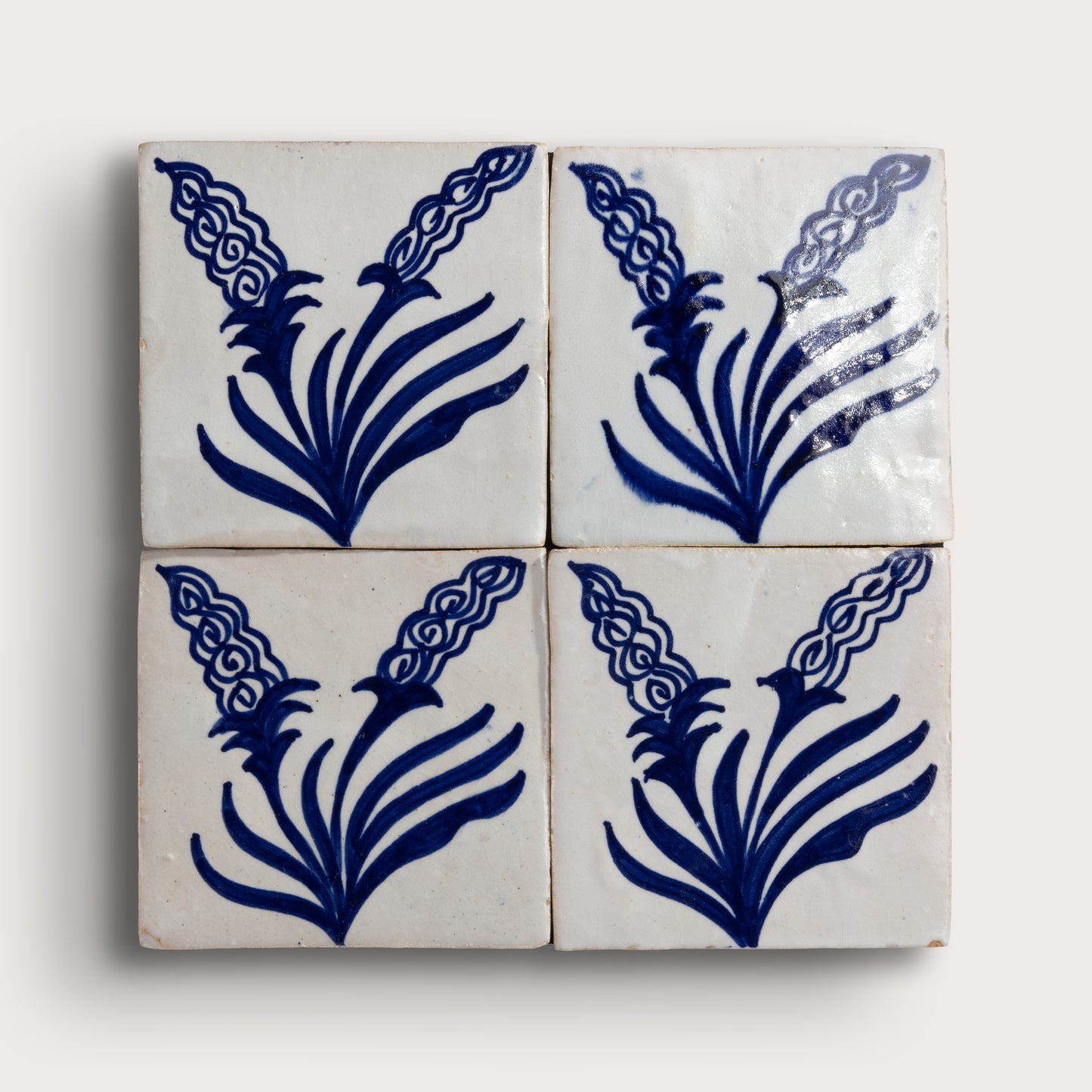Four square zellige tiles arranged in a 2x2 grid, each decorated with a dark blue floral or leaf-like motif on an off-white background, photographed against a light neutral backdrop.