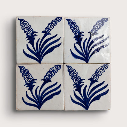 Four square zellige tiles arranged in a 2x2 grid, each decorated with a dark blue floral or leaf-like motif on an off-white background, photographed against a light neutral backdrop.