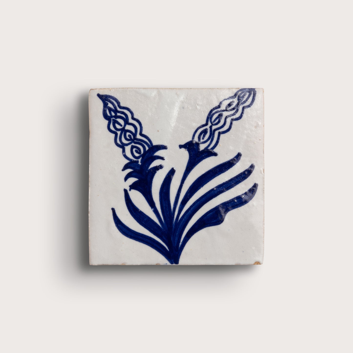 square zellige tile with a white glaze and a blue hand-painted floral pattern, on a light beige background.