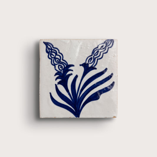 square zellige tile with a white glaze and a blue hand-painted floral pattern, on a light beige background.