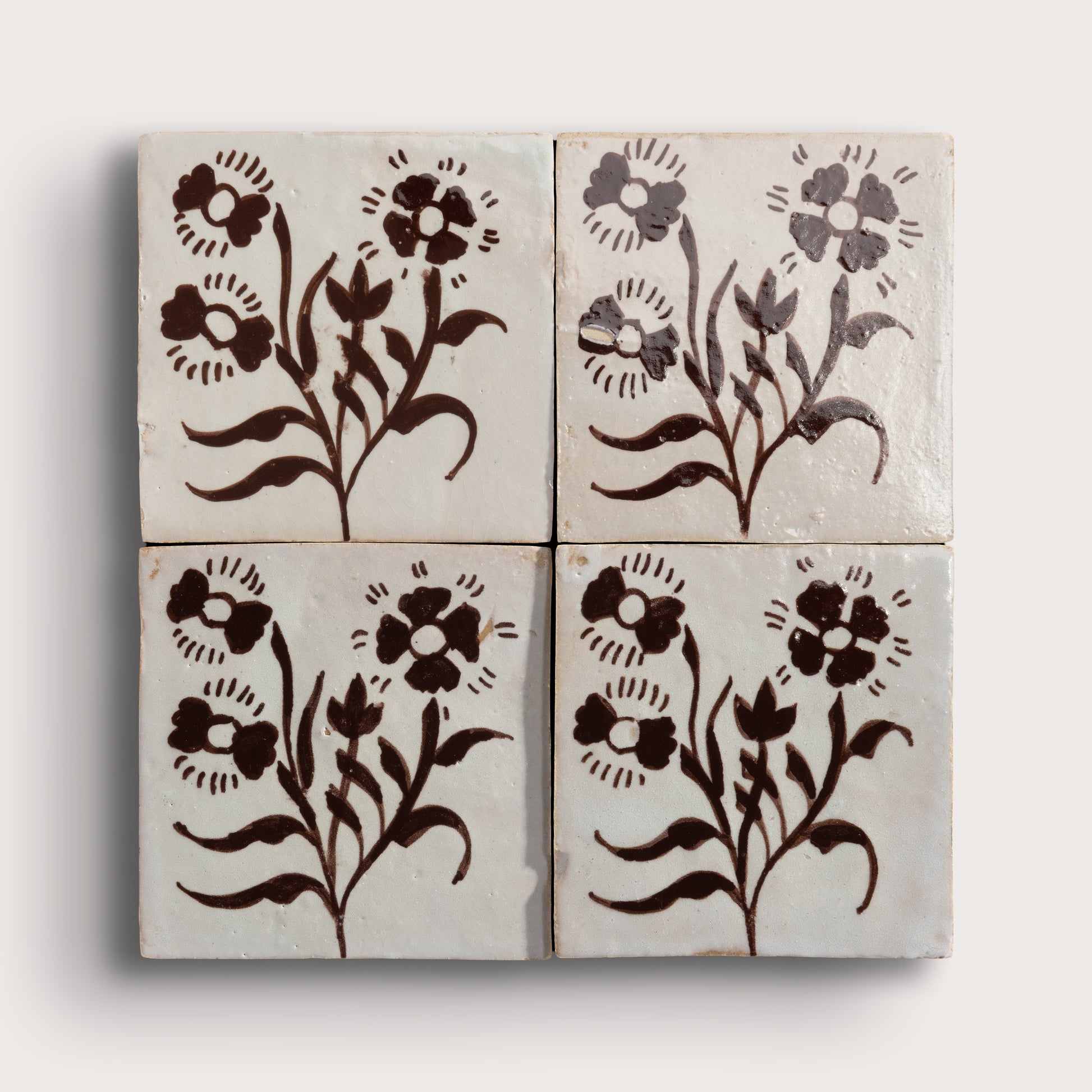 Four square zellige tiles arranged in a 2x2 grid, each featuring a dark brown floral motif on a warm off-white background, photographed against a light neutral wall.