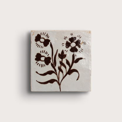 square zellige tile, featuring a dark brown floral motif on a warm off-white background, photographed against a light neutral wall.