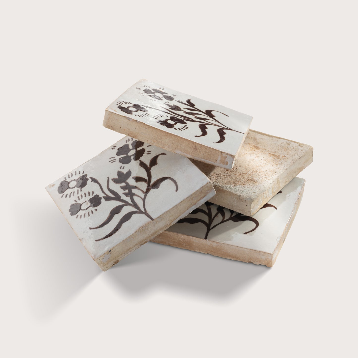 Four square zellige tiles stacked, each featuring a dark brown floral motif on a warm off-white background, photographed against a light neutral wall.