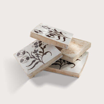 Four square zellige tiles stacked, each featuring a dark brown floral motif on a warm off-white background, photographed against a light neutral wall.