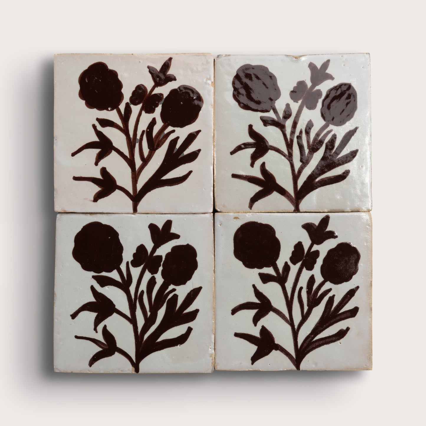 Four square zellige tiles arranged in a 2x2 grid, each featuring a dark brown botanical floral motif on a warm off-white glazed background, photographed against a light neutral wall.