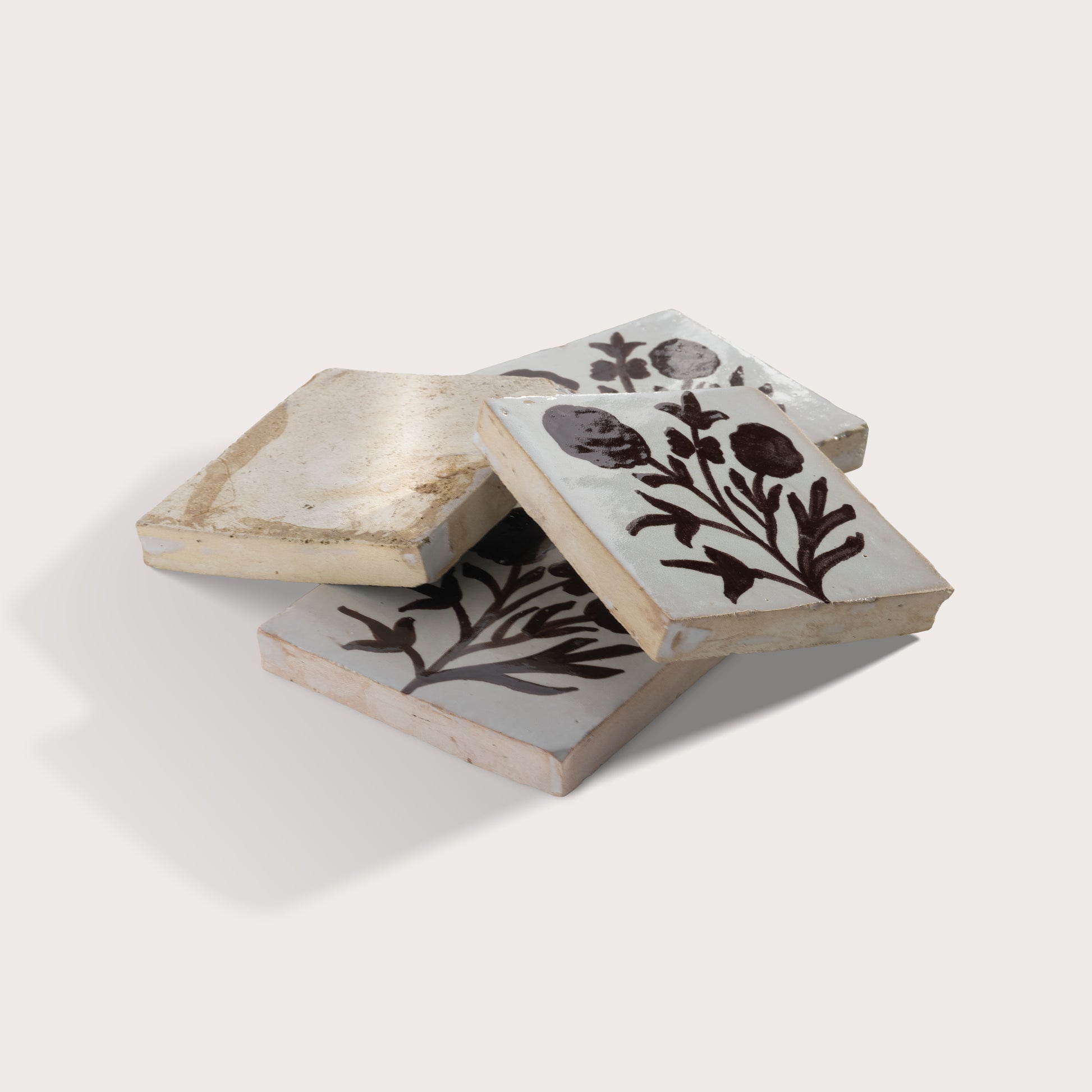 Three square zellige tiles with a rustic hand-painted botanical design in dark brown on a light gray background, arranged in a stack on a light beige studio background.