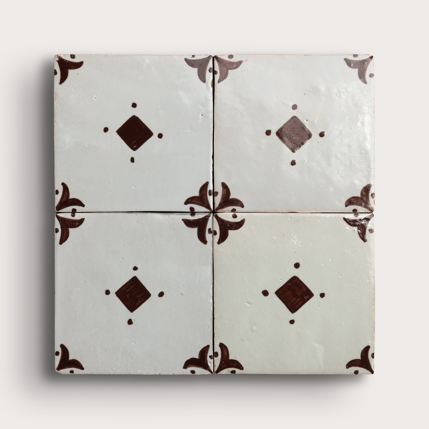 Four square zellige tiles arranged in a 2x2 grid, featuring a glossy off-white glaze with dark brown decorative floral corner motifs and diamond accents, set against a light neutral background.
