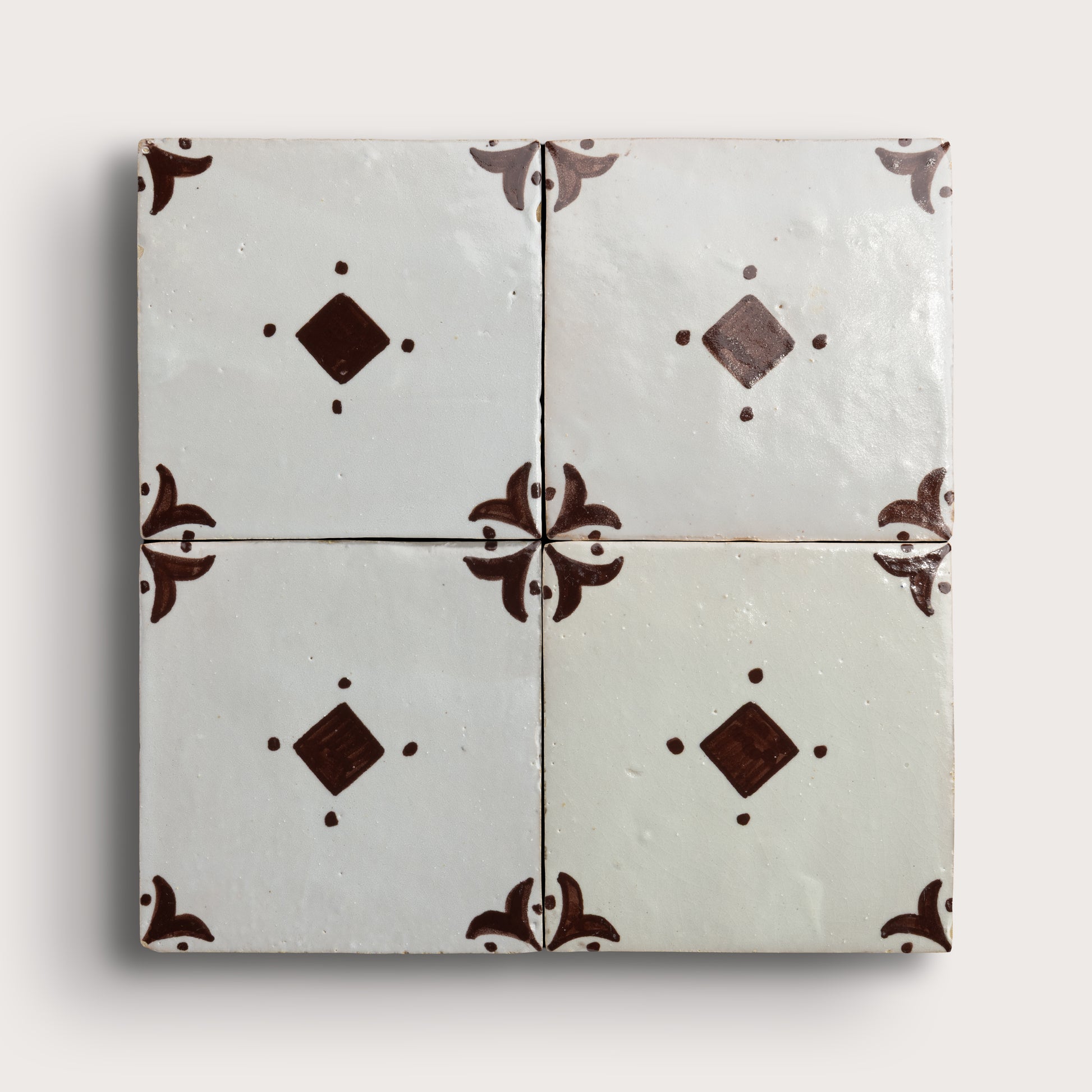Four square zellige tiles arranged in a 2x2 grid, featuring a glossy off-white glaze with dark brown decorative floral corner motifs and diamond accents, set against a light neutral background.