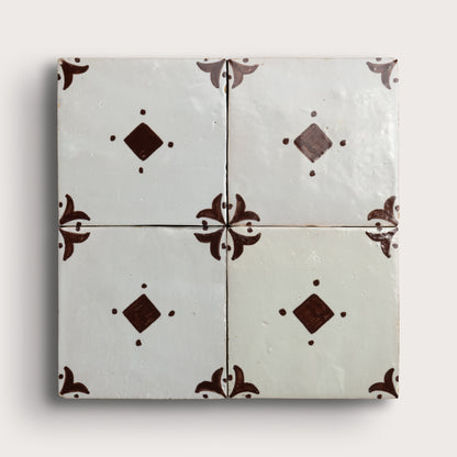 Four square zellige tiles arranged in a 2x2 grid, featuring a glossy off-white glaze with dark brown decorative floral corner motifs and diamond accents, set against a light neutral background.