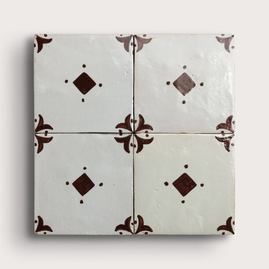 Four square zellige tiles arranged in a 2x2 grid, featuring a glossy off-white glaze with dark brown decorative floral corner motifs and diamond accents, set against a light neutral background.