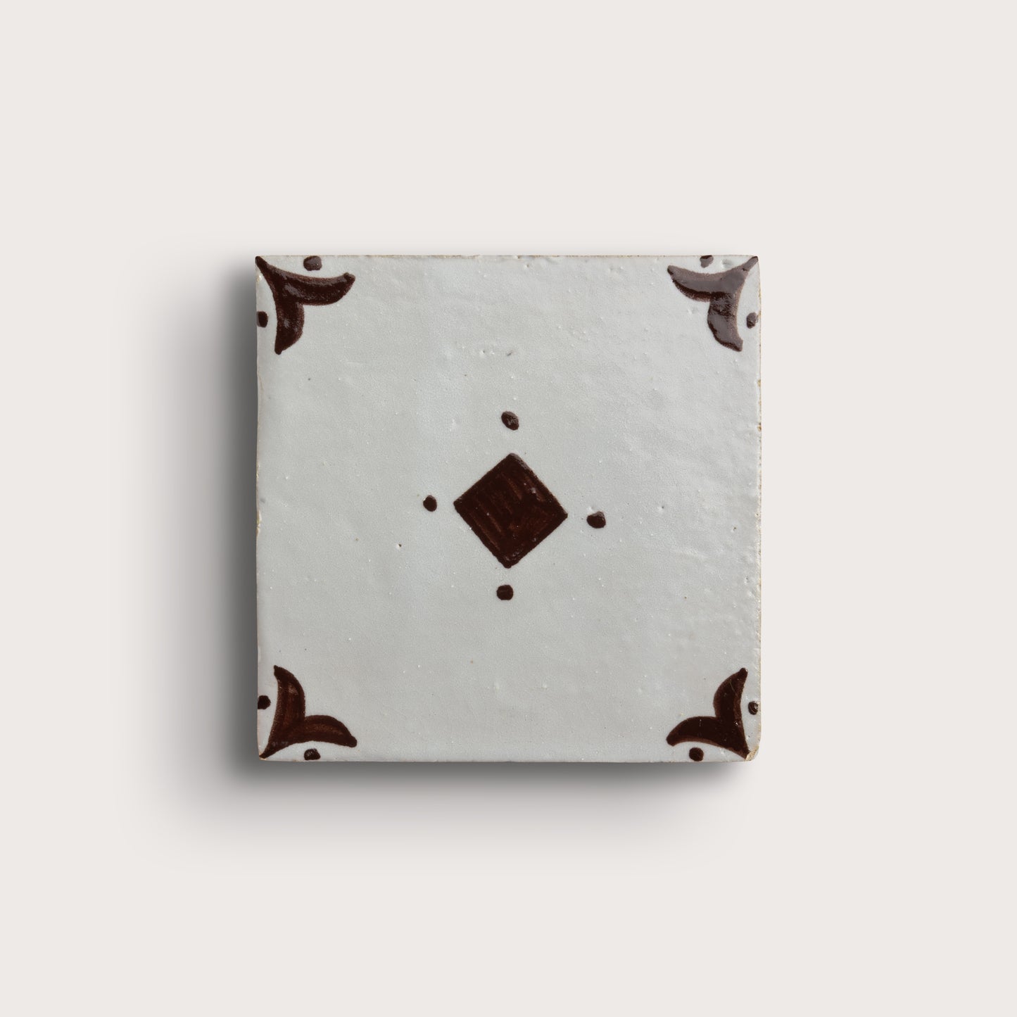 A square zellige tile with a light gray glaze, featuring a central dark brown diamond motif and matching dark brown corner flourishes, shown on a light beige background.
