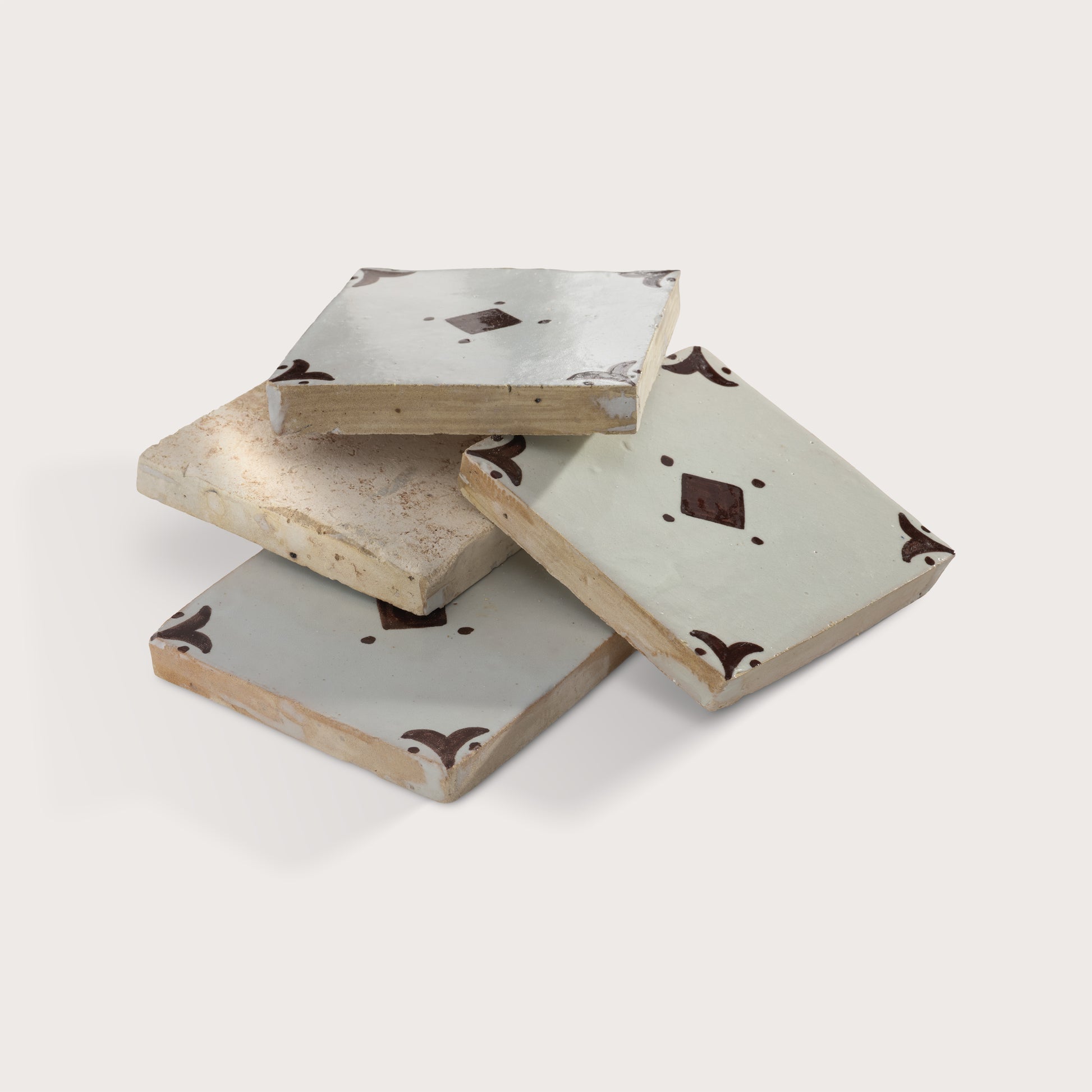 Stacked square zellige tiles with a light gray glaze, featuring a central dark brown diamond motif and matching dark brown corner flourishes, shown on a light beige background.