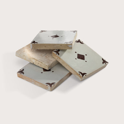 Stacked square zellige tiles with a light gray glaze, featuring a central dark brown diamond motif and matching dark brown corner flourishes, shown on a light beige background.