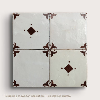 Four square zellige tiles arranged in a 2x2 grid, featuring a glossy off-white glaze with dark brown decorative floral corner motifs and diamond accents, set against a light neutral background.