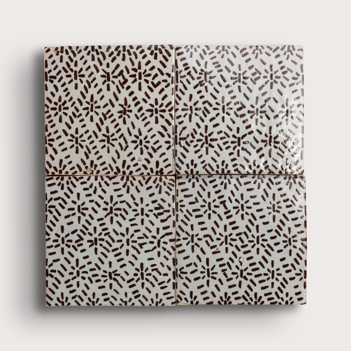 Four square zellige tiles in a grid pattern on a light gray background. Two tiles feature a brown hand-painted abstract brushstroke pattern on a white glaze, while the bottom tile shows a textured, unglazed beige clay surface.