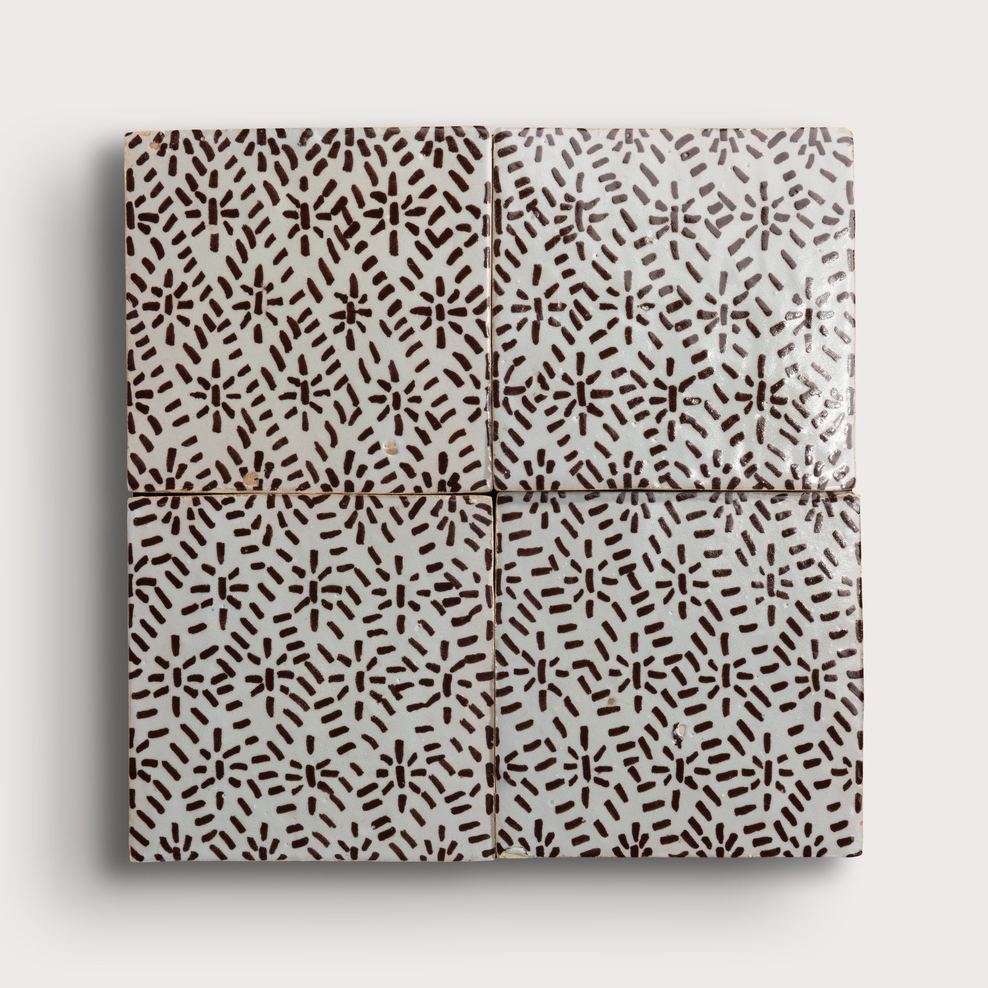 Four square zellige tiles in a grid pattern on a light gray background. Two tiles feature a brown hand-painted abstract brushstroke pattern on a white glaze, while the bottom tile shows a textured, unglazed beige clay surface.