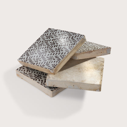 Four square zellige tiles stacked and overlapping on a light gray background. Two tiles feature a brown hand-painted abstract brushstroke pattern on a white glaze, while the bottom tile shows a textured, unglazed beige clay surface.