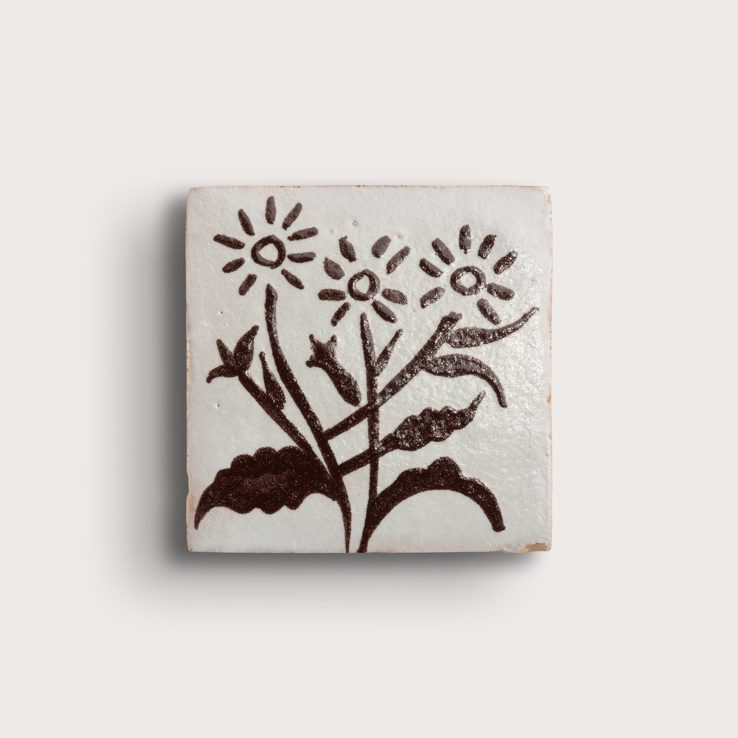 A square zellige tile featuring a hand-painted dark brown floral design on a glossy off-white glaze, set against a plain light beige background.