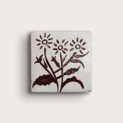 A square zellige tile featuring a hand-painted dark brown floral design on a glossy off-white glaze, set against a plain light beige background.