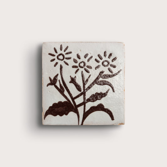 A square zellige tile featuring a hand-painted dark brown floral design on a glossy off-white glaze, set against a plain light beige background.