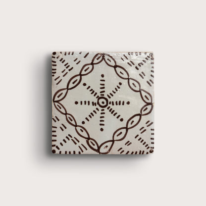 square zellige tile with a handpainted brown pattern shot on a light gray background.
