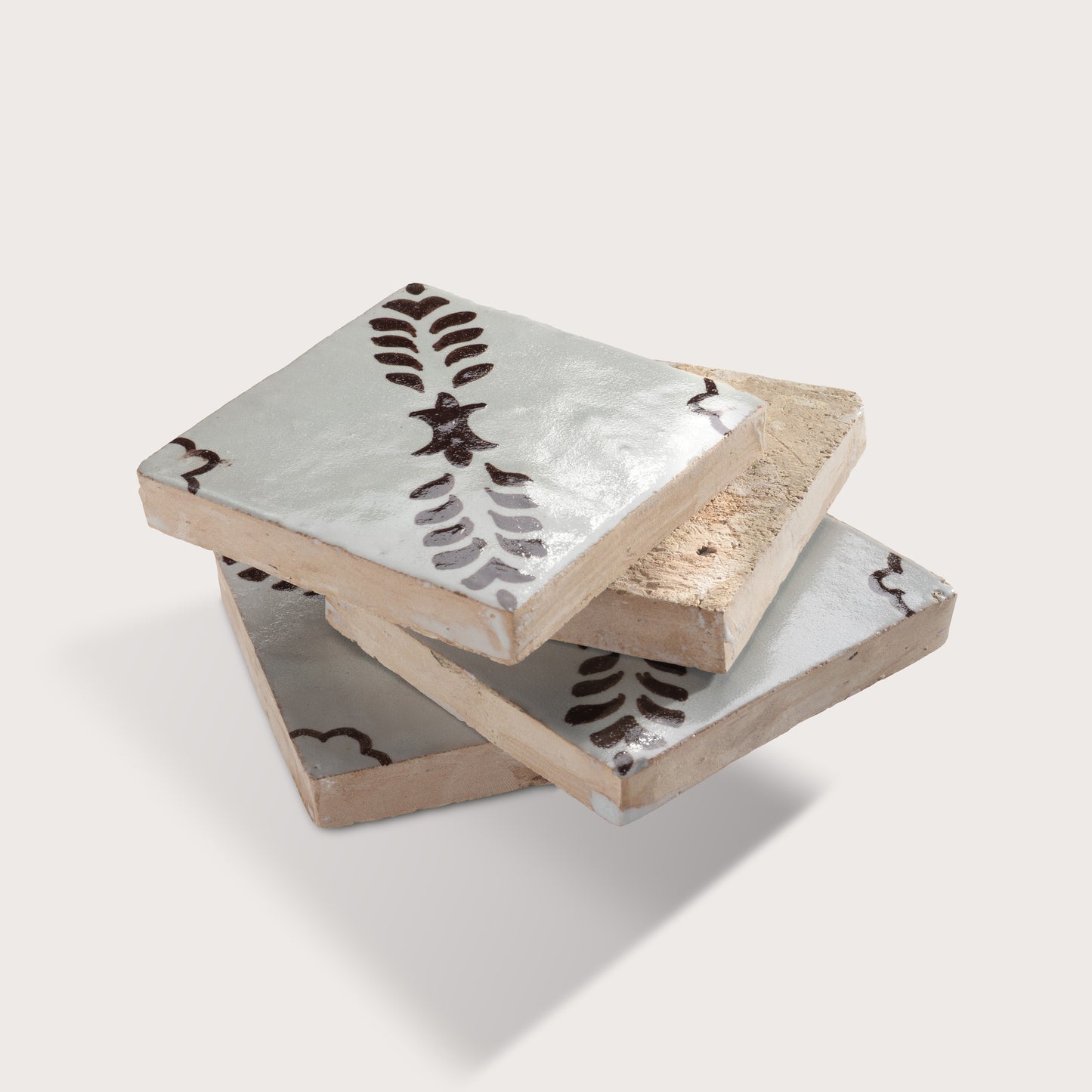Four square zellige tiles stacked, featuring a glossy off-white glaze with a dark brown hand-painted botanical pattern of leafy stems and star-like motifs.