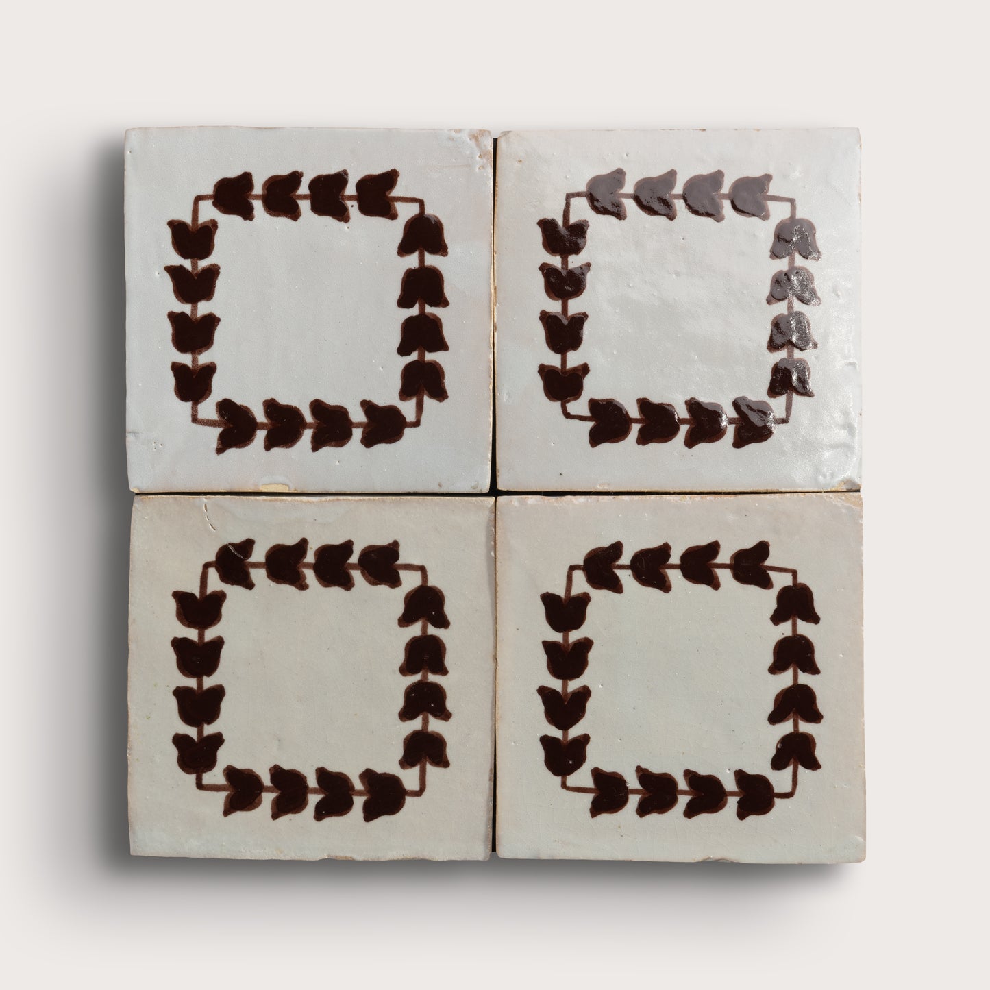 Four square zellige tiles arranged in a 2x2 grid on a light neutral background. Two tiles feature a dark brown leafy border pattern, and two are plain off-white with brown corner accents.