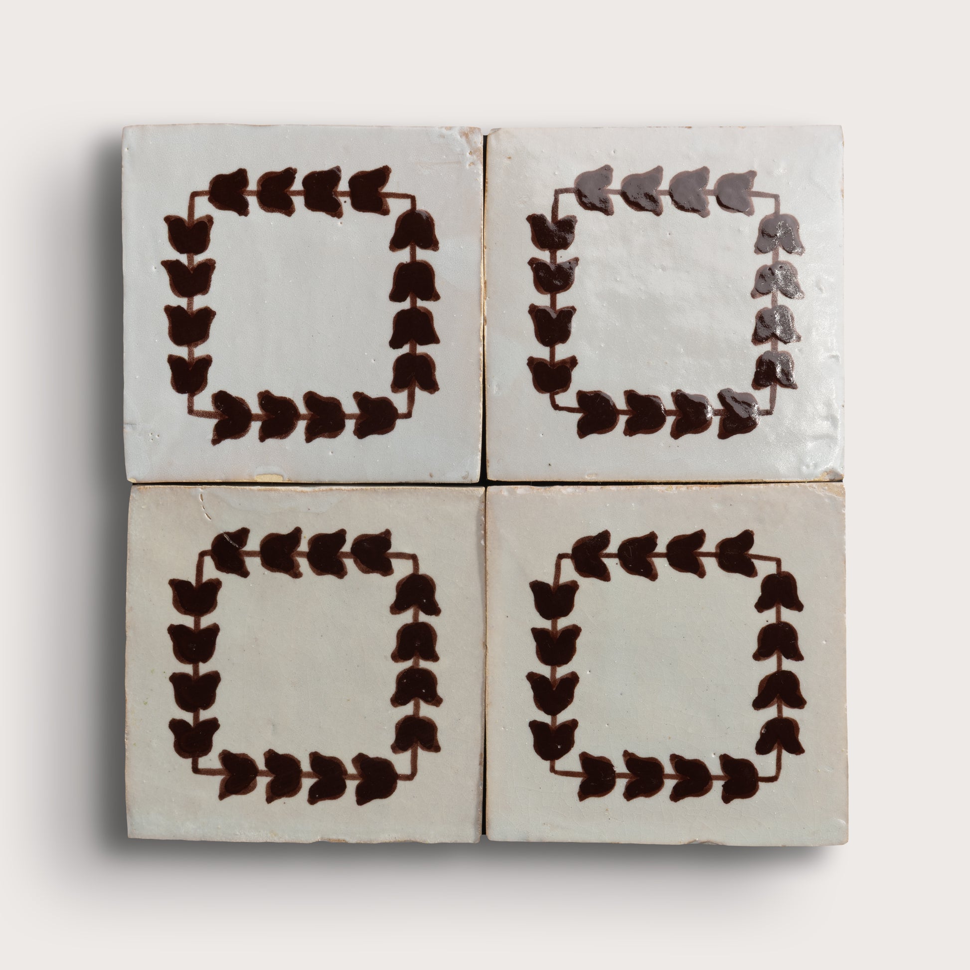 Four square zellige tiles arranged in a 2x2 grid on a light neutral background. Two tiles feature a dark brown leafy border pattern, and two are plain off-white with brown corner accents.