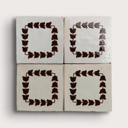 Four square zellige tiles arranged in a 2x2 grid on a light neutral background. Two tiles feature a dark brown leafy border pattern, and two are plain off-white with brown corner accents.