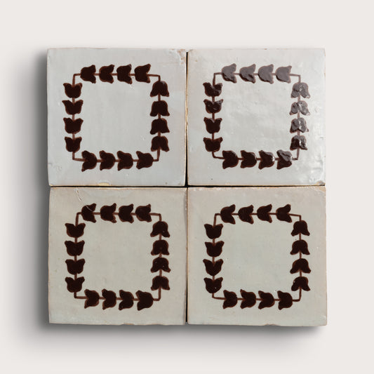 Four square zellige tiles arranged in a 2x2 grid on a light neutral background. Two tiles feature a dark brown leafy border pattern, and two are plain off-white with brown corner accents.